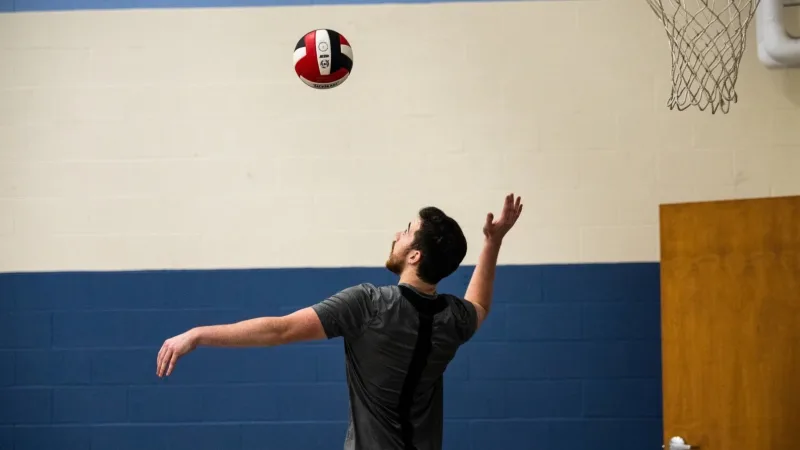 Man serving a volleyball overhead in a school gym, players at the net behind